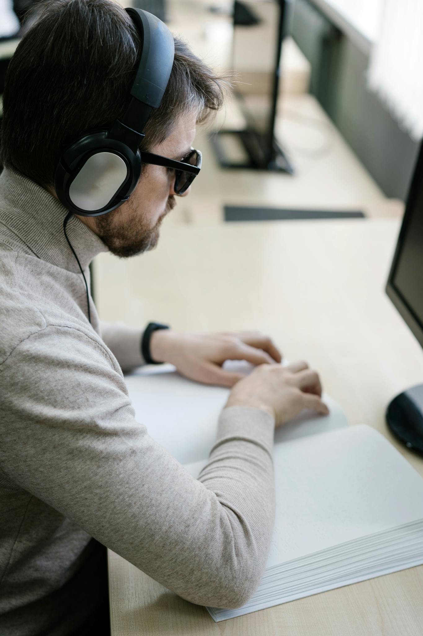 Adult man using braille and headphones at a desk in an office setting, focusing on accessibility.